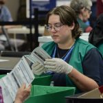 Election workers verify mail ballots in a counting facility, wearing gloves and green vests. — USPS mail ballot rules
