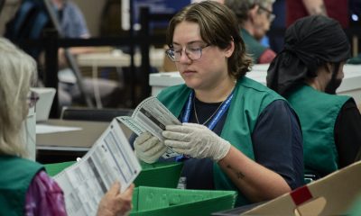 Election workers verify mail ballots in a counting facility, wearing gloves and green vests. — USPS mail ballot rules