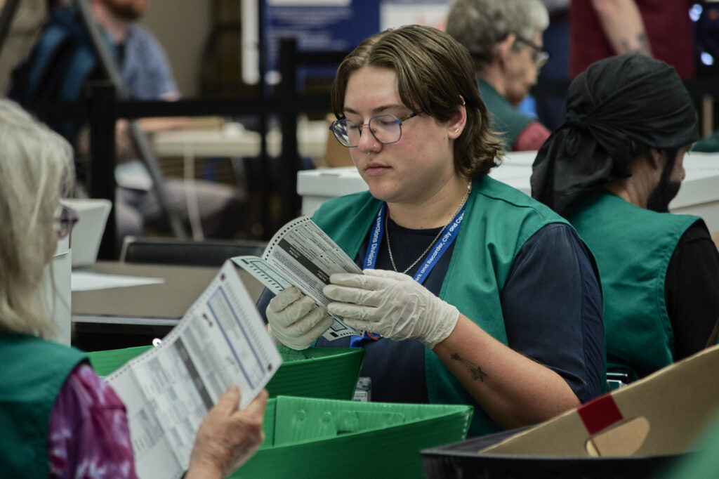 Election workers verify mail ballots in a counting facility, wearing gloves and green vests. — USPS mail ballot rules