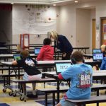 Students in a school cafeteria using tablets while wearing headphones, with a teacher assisting them. — AI edtech spending
