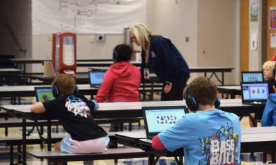 Students in a school cafeteria using tablets while wearing headphones, with a teacher assisting them. — AI edtech spending