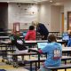 Students in a school cafeteria using tablets while wearing headphones, with a teacher assisting them. — AI edtech spending