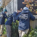 FBI agents in jackets examining a bush near a fence, highlighting crime investigation efforts. — FBI crime report 2025