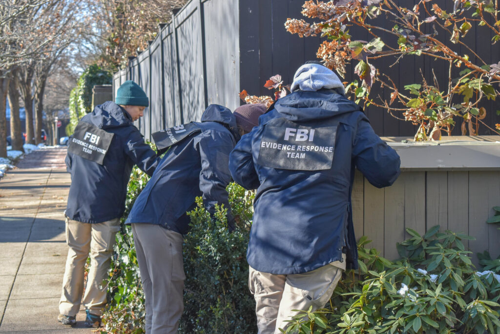 FBI agents in jackets examining a bush near a fence, highlighting crime investigation efforts. — FBI crime report 2025