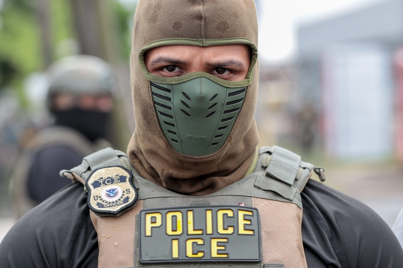 An ICE officer in tactical gear with a mask, displaying a badge and the word 'POLICE' on his — ICE monthly arrests record