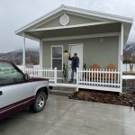 A man stands on the porch of a small house, holding a lamp, with a truck parked nearby; a sign of veteran housing assistance.