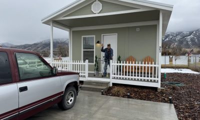 A man stands on the porch of a small house, holding a lamp, with a truck parked nearby; a sign of veteran housing assistance.