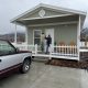 A man stands on the porch of a small house, holding a lamp, with a truck parked nearby; a sign of veteran housing assistance.