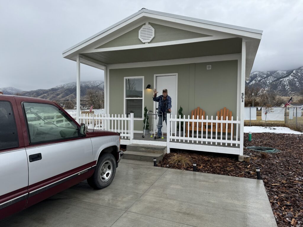 A man stands on the porch of a small house, holding a lamp, with a truck parked nearby; a sign of veteran housing assistance.