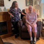 A caregiver stands next to an elderly woman sitting in a recliner in a cozy living room, highlight — in home caregiver pay