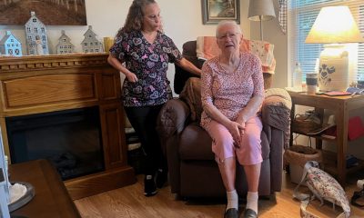 A caregiver stands next to an elderly woman sitting in a recliner in a cozy living room, highlight — in home caregiver pay