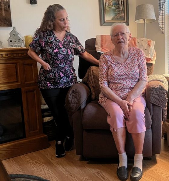 A caregiver stands next to an elderly woman sitting in a recliner in a cozy living room, highlight — in home caregiver pay