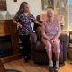 A caregiver stands next to an elderly woman sitting in a recliner in a cozy living room, highlight — in home caregiver pay