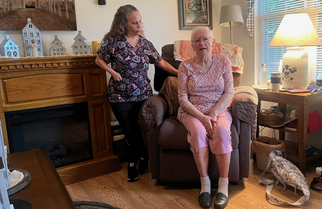 A caregiver stands next to an elderly woman sitting in a recliner in a cozy living room, highlight — in home caregiver pay