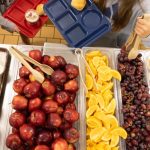 A school cafeteria display with trays of apples, oranges, grapes, and desserts, highlighting — SNAP cuts free school meals
