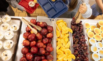A school cafeteria display with trays of apples, oranges, grapes, and desserts, highlighting — SNAP cuts free school meals