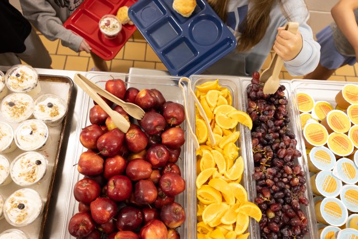 A school cafeteria display with trays of apples, oranges, grapes, and desserts, highlighting — SNAP cuts free school meals