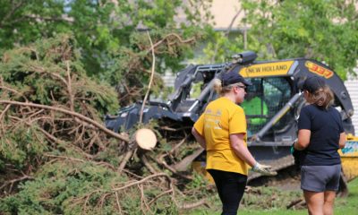 Two women discuss cleanup efforts near a tree debris pile and heavy machinery after a storm, highlighting FEMA disaster aid.