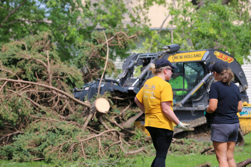 Two women discuss cleanup efforts near a tree debris pile and heavy machinery after a storm, highlighting FEMA disaster aid.