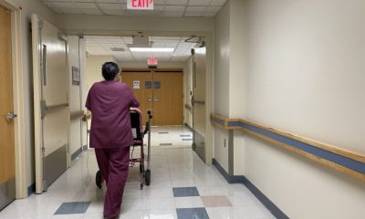 A healthcare worker in scrubs pushing a patient in a walker down a hospital corridor, highlighting Medicaid payment cuts i…