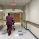 A healthcare worker in scrubs pushing a patient in a walker down a hospital corridor, highlighting Medicaid payment cuts i…