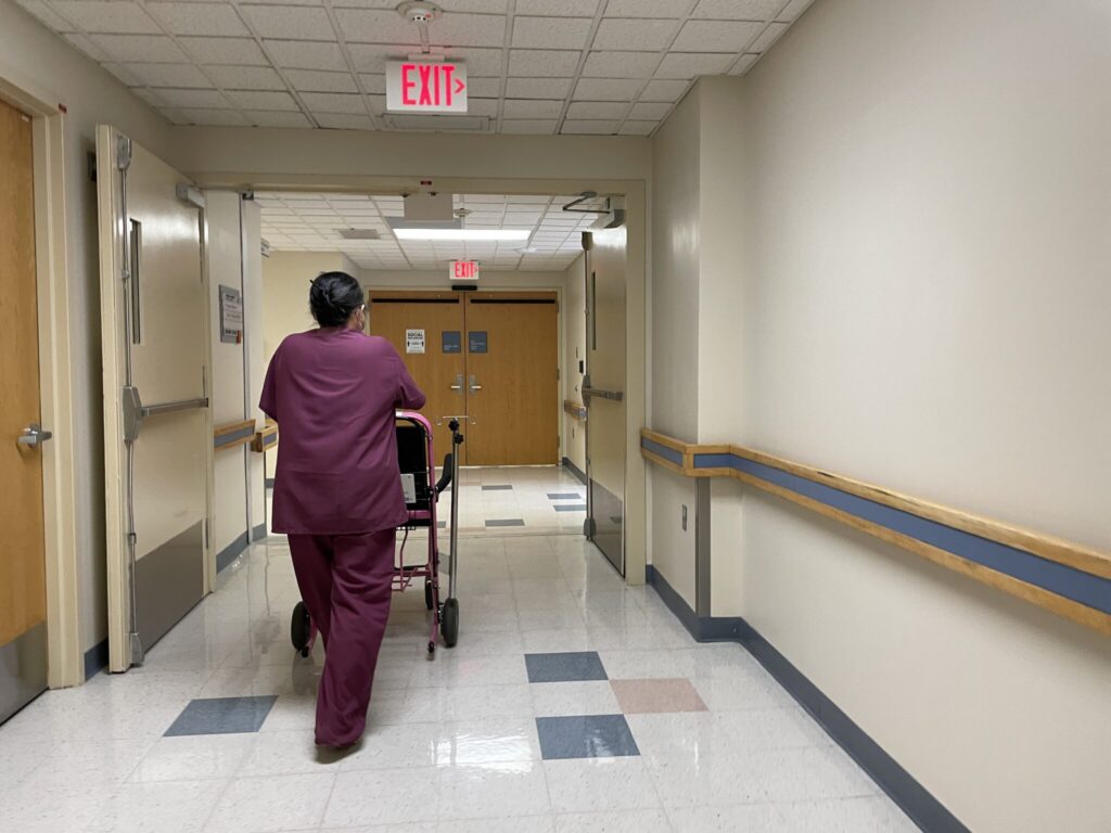 A healthcare worker in scrubs pushing a patient in a walker down a hospital corridor, highlighting Medicaid payment cuts i…