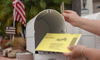 A hand placing a yellow mail-in ballot into a white mailbox with an American flag in t — USPS mail-in voting whistleblower