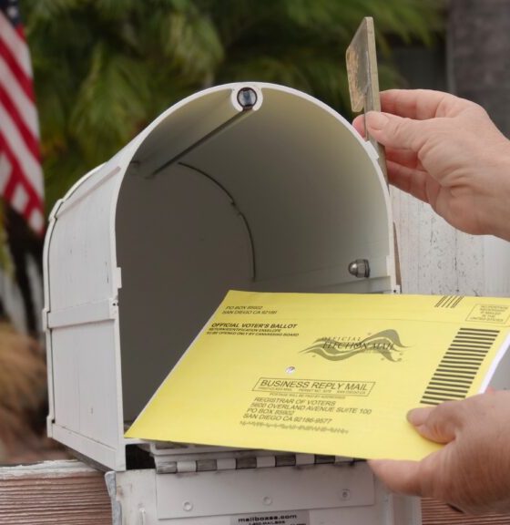 A hand placing a yellow mail-in ballot into a white mailbox with an American flag in t — USPS mail-in voting whistleblower