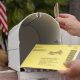 A hand placing a yellow mail-in ballot into a white mailbox with an American flag in t — USPS mail-in voting whistleblower
