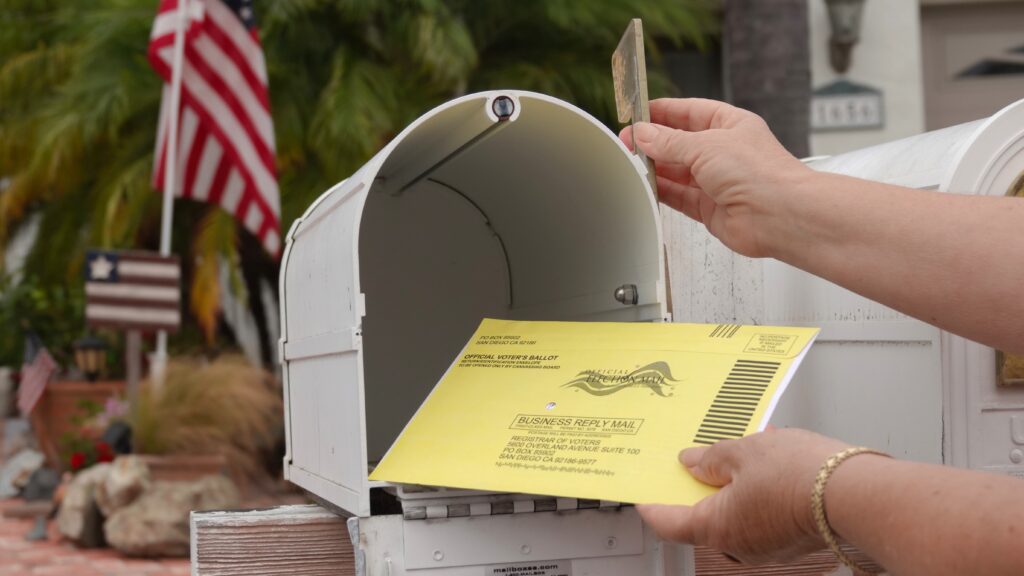 A hand placing a yellow mail-in ballot into a white mailbox with an American flag in t — USPS mail-in voting whistleblower