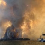 Smoke and flames rise from a wildfire near the Colville Reservation, with a boat in the f — Colville Reservation Wildfires