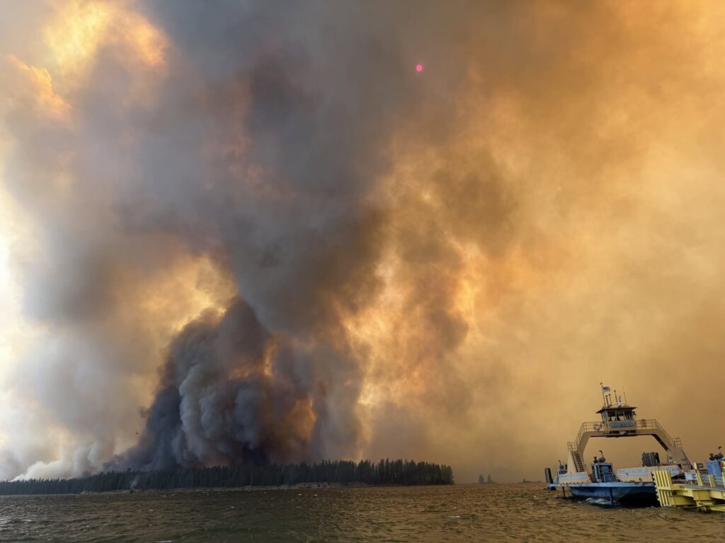 Smoke and flames rise from a wildfire near the Colville Reservation, with a boat in the f — Colville Reservation Wildfires
