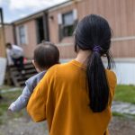 A woman holding a baby walks towards a mobile home, symbolizing the challenges faced by mixed-sta — new public charge rule