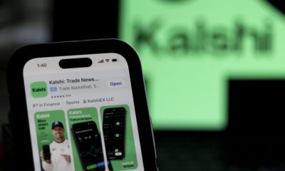 Utah Sues Kalshi: a phone displays the Kalshi trading app against a blurred green Kalshi sign.