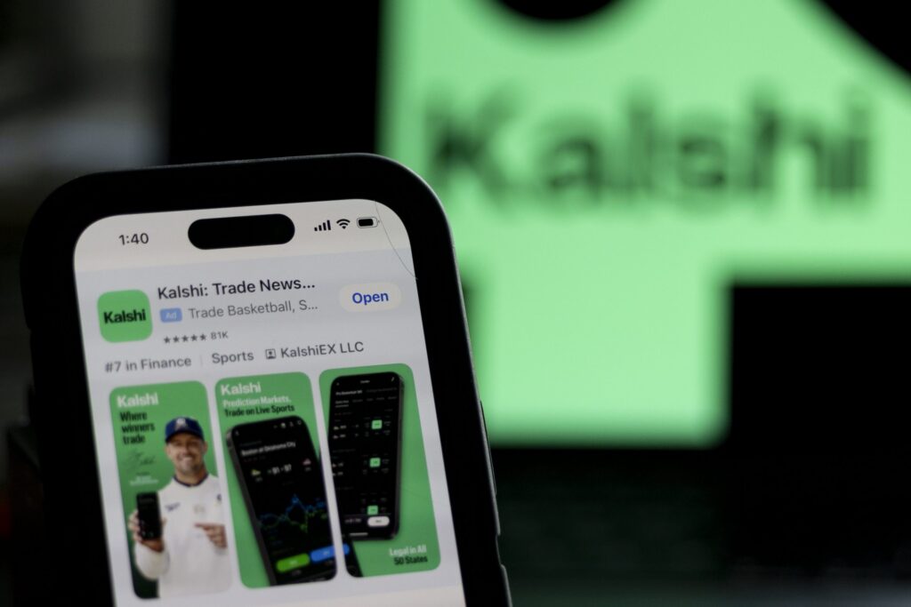 Utah Sues Kalshi: a phone displays the Kalshi trading app against a blurred green Kalshi sign.