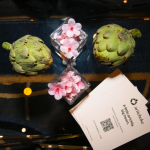Artichokes, pink flowers, and an Artichoke Credit Union business card arranged on a reflective table.