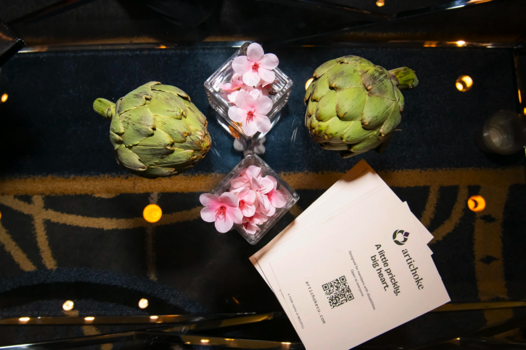 Artichokes, pink flowers, and an Artichoke Credit Union business card arranged on a reflective table.