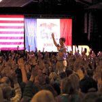 A crowd raises their hands in support during a political rally, with flags in the background — Democrats governor majority