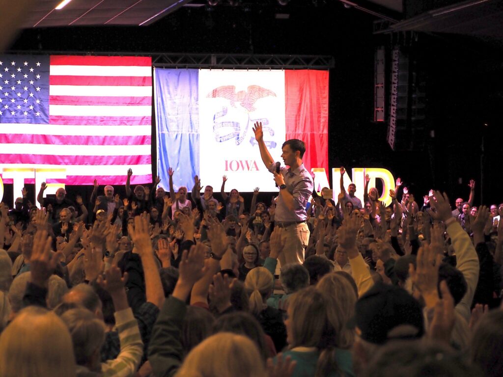 A crowd raises their hands in support during a political rally, with flags in the background — Democrats governor majority