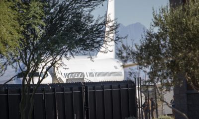 Trump deportation statistics: a white aircraft behind a tall border fence, with people visible nearby.