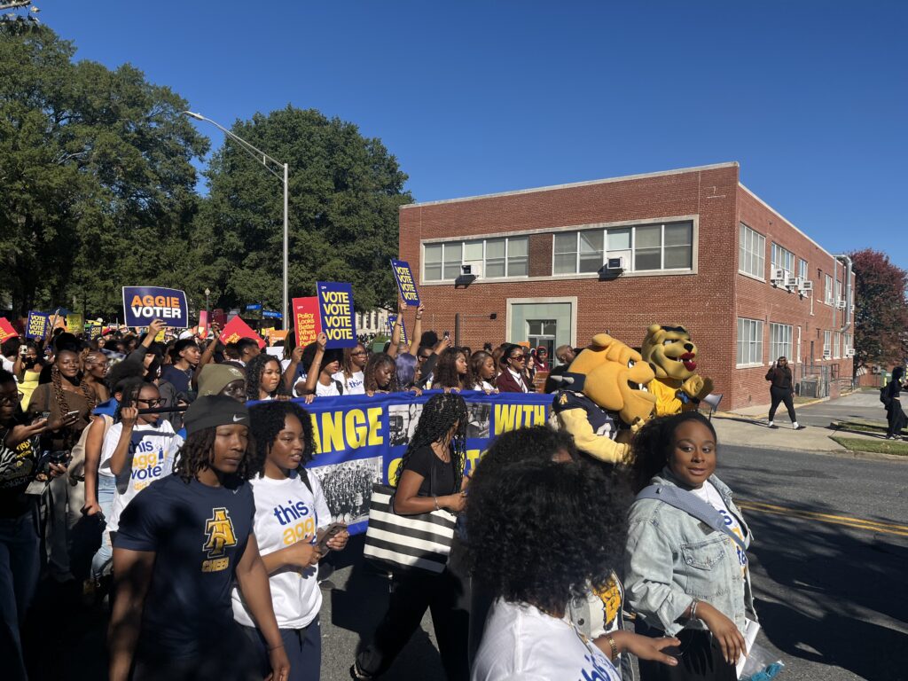 NC A&T campus polling site march with students carrying voter signs and university mascots on a sunny street