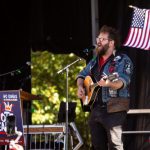 Musician performing at a No Kings early voting day of action event, with an American flag in the background.