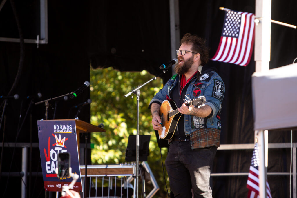 Musician performing at a No Kings early voting day of action event, with an American flag in the background.