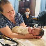 A midwife cradles a newborn beside a masked caregiver; states without birth centers face gaps in maternal care.