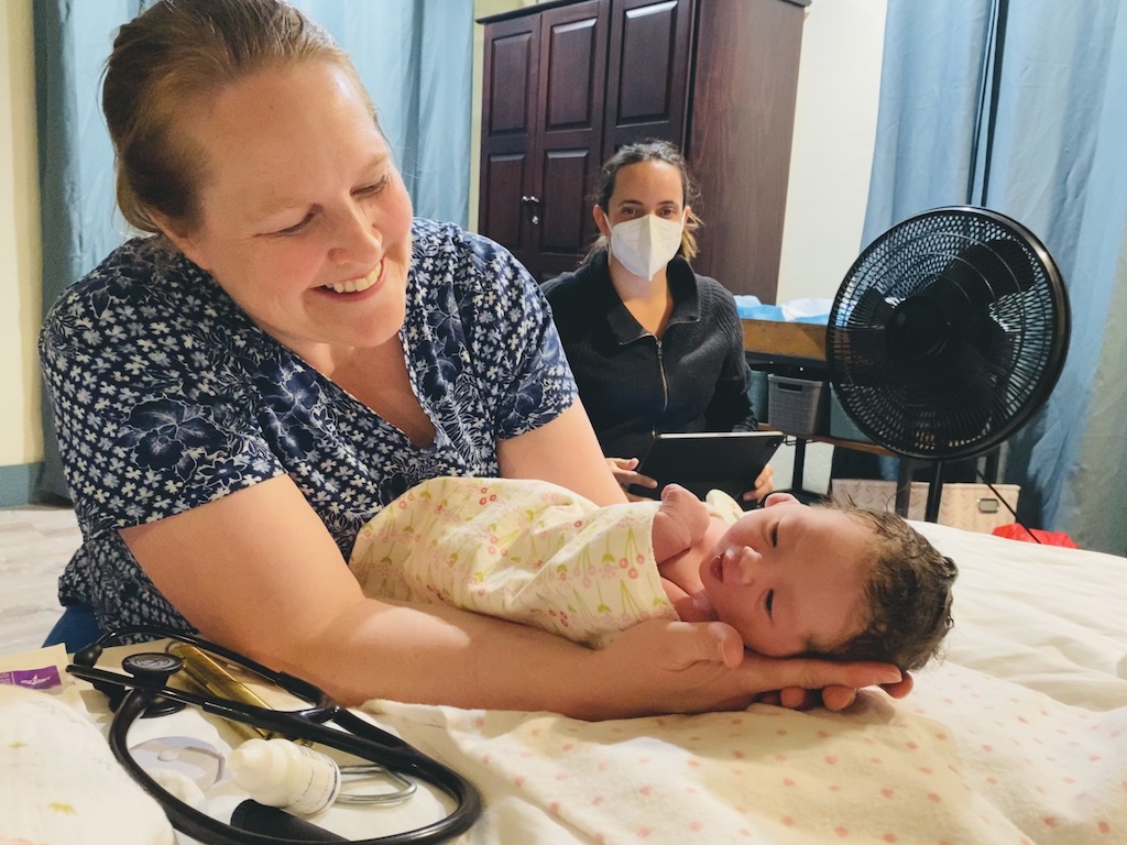 A midwife cradles a newborn beside a masked caregiver; states without birth centers face gaps in maternal care.