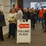 Voters queue inside a public building beside a “Vote Here” polling place sign, amid Trump DOJ election lawsuits.