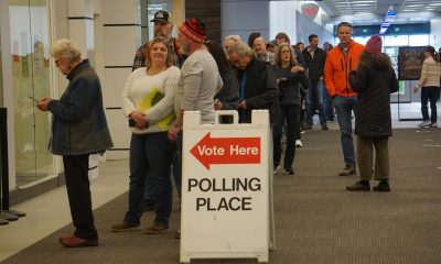 Voters queue inside a public building beside a “Vote Here” polling place sign, amid Trump DOJ election lawsuits.