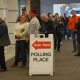 Voters queue inside a public building beside a “Vote Here” polling place sign, amid Trump DOJ election lawsuits.