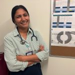 Smiling female doctor with a stethoscope, standing in front of a bulletin boa — Trump Immigration Policies Foreign Doctors