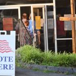 DOJ election monitors 2026 story: voters enter a polling place beside a prominent “Vote Here” sign.